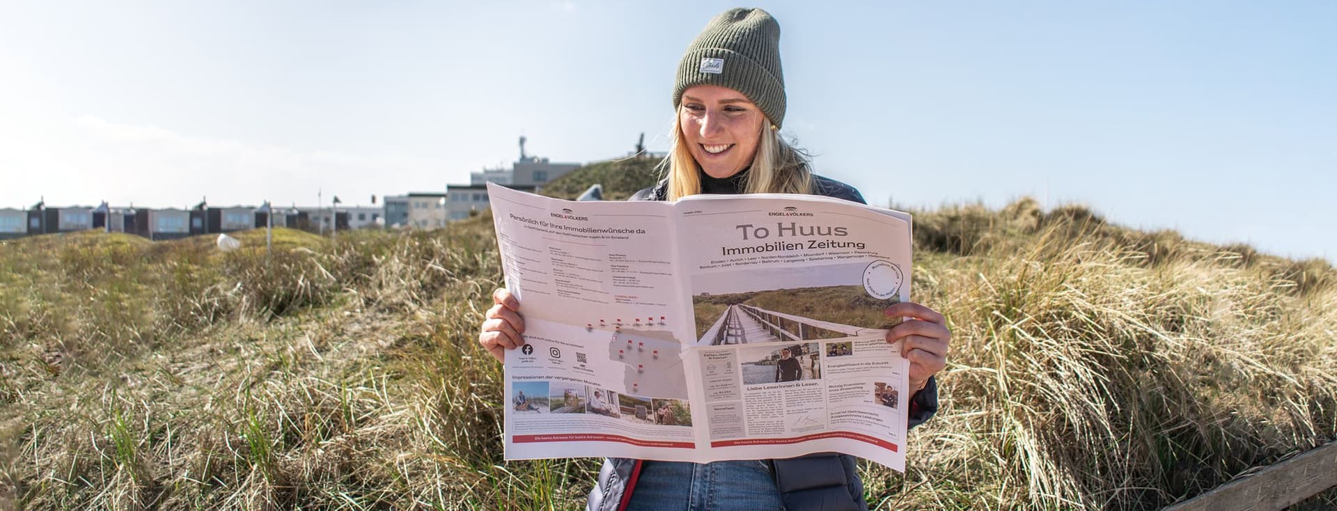 Frau liest die Engel & Völkers Immobilienzeitung „To Huus“ in den Dünen Ostfrieslands bei Sonnenschein und frischer Nordseeluft