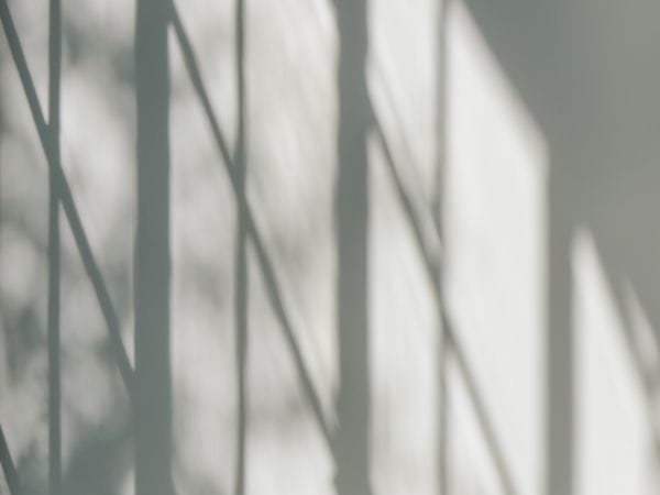 Gelassene Sitzecke mit einem weißen Sofa mit grauen Kissen und einem runden Couchtisch aus Holz. Dramatische Schatten, die von den Fensterrahmen geworfen werden, erzeugen geometrische Muster an der hellgrauen Wand und verleihen dem minimalistischen Raum Tiefe und visuelles Interesse in einer ruhigen, monochromen Palette.