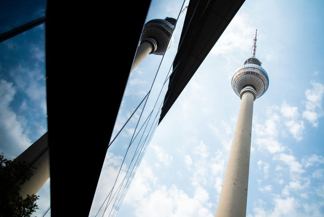 Low-angle view of the TV tower with antenna against a blue sky, mirrored in the angled glass facade of a nearby building