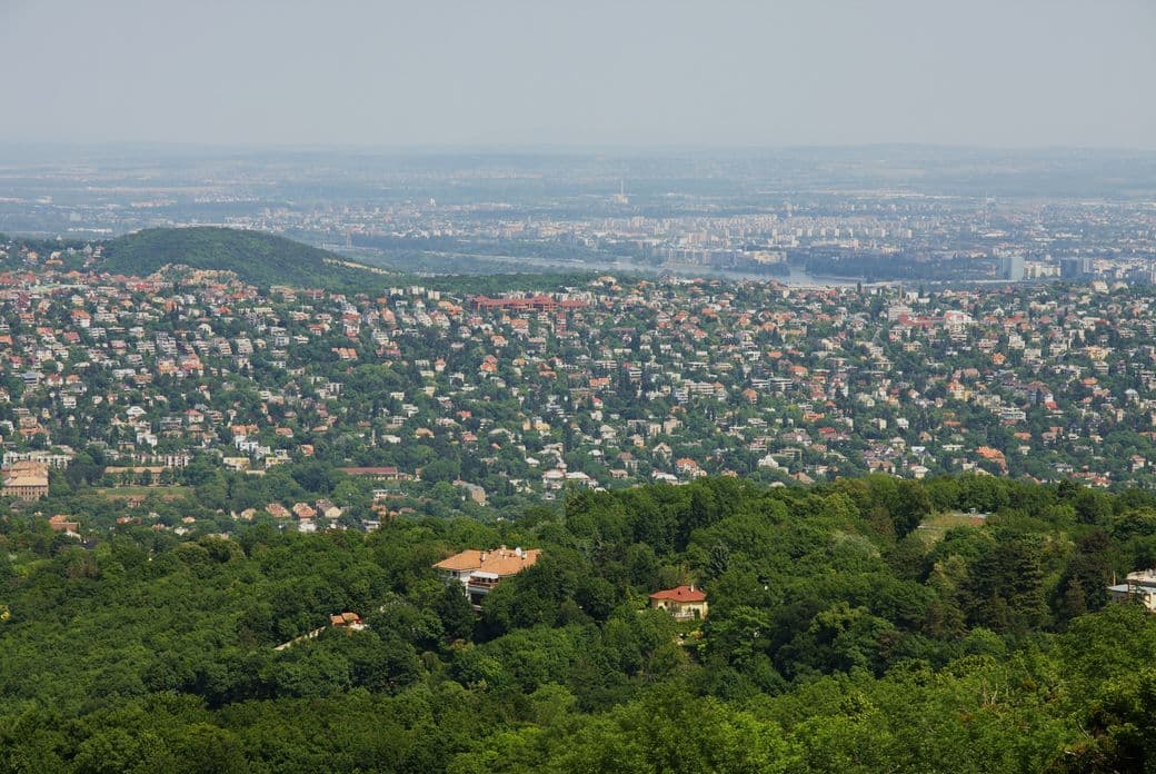 Aerial view of a lush, green landscape with scattered houses, hills, and a distant city skyline under a clear sky.