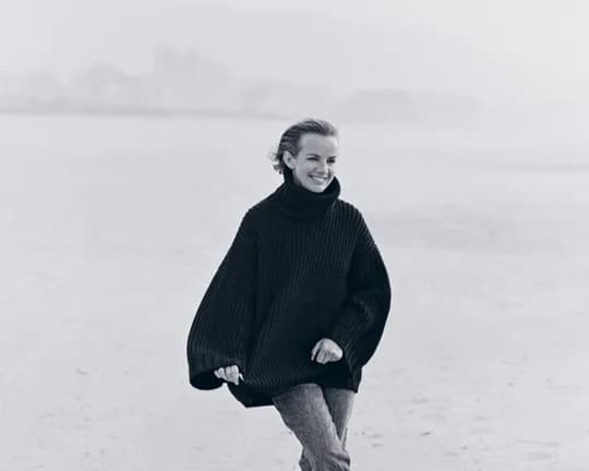 A person in a chunky dark sweater walks along a misty beach, smiling, with water and distant shoreline in the background.