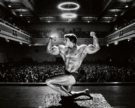 Black-and-white photo of a muscular bodybuilder posing on a stage, flexing arms before a large theater audience.
