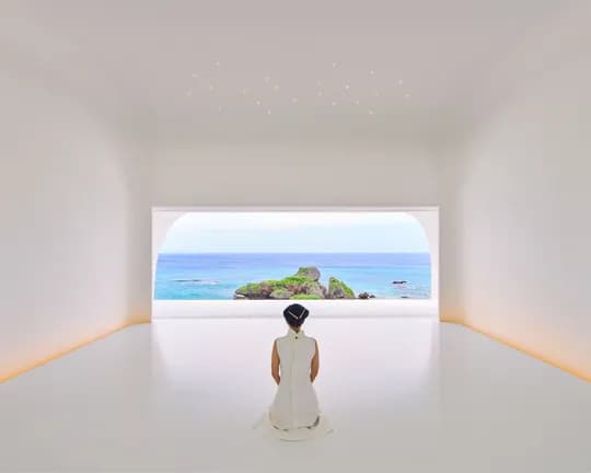 A woman in a white dress kneels in a bright, minimalist room, facing an arched window onto a blue sea and rocky island.