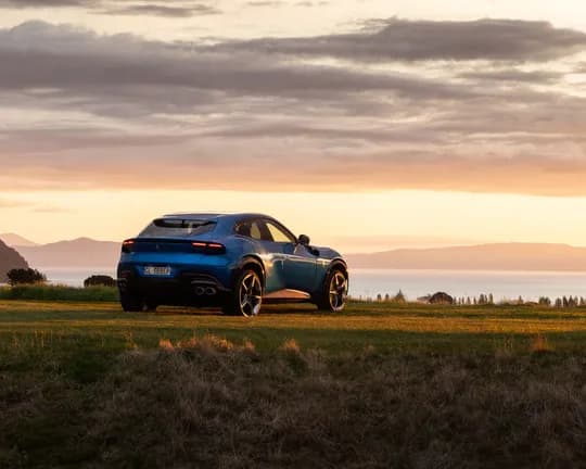 Blue SUV on a grassy hillside at sunset, overlooking a lake with distant mountains and a warm orange sky.