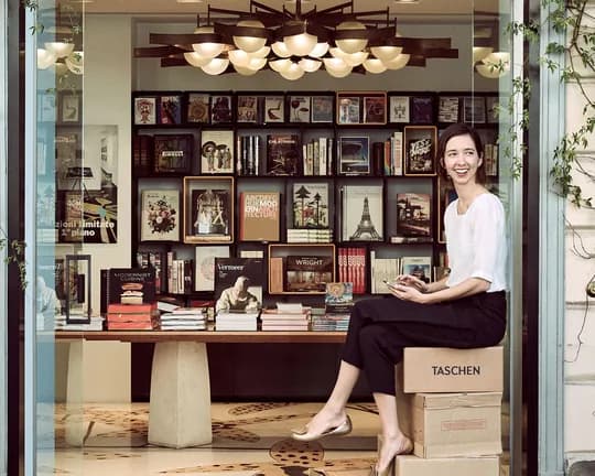 Smiling woman sits on a TASCHEN box at the doorway of a bookstore, with shelves of books and framed art visible inside.