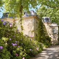 Sunlit garden with purple rhododendrons around a beige historic building; a gravel path winds past tall trees.
