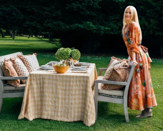 A woman in an orange floral dress stands beside a table with a yellow checkered cloth, set for an outdoor meal on a grassy lawn with cushioned chairs.