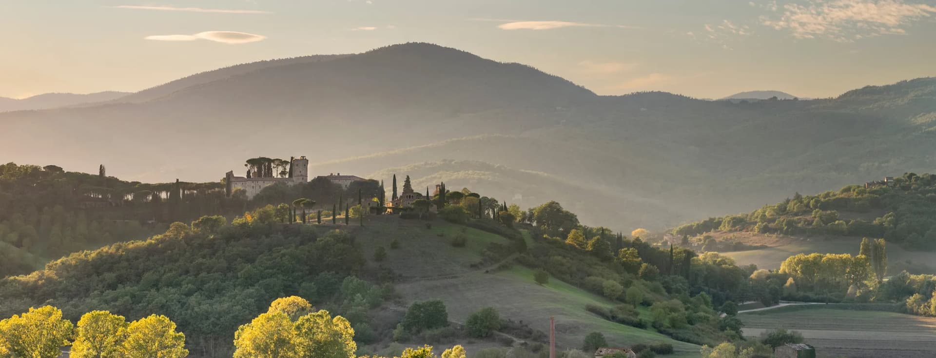 Die goldenen Hügel der Toskana im Morgengrauen, eine Burg auf einem Hügel mit Zypressen, nebelverhangene Täler und leuchtend bunte Herbstbäume im Vordergrund.