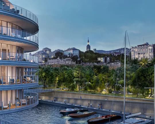 Curved glass apartment building along a marina at dusk, with lit balconies, nearby boats, and a city skyline with a tall church steeple.