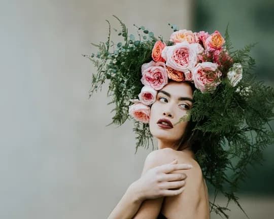 Woman wearing a large floral crown of pink and peach roses with greenery, bare shoulders, gazing to the side.