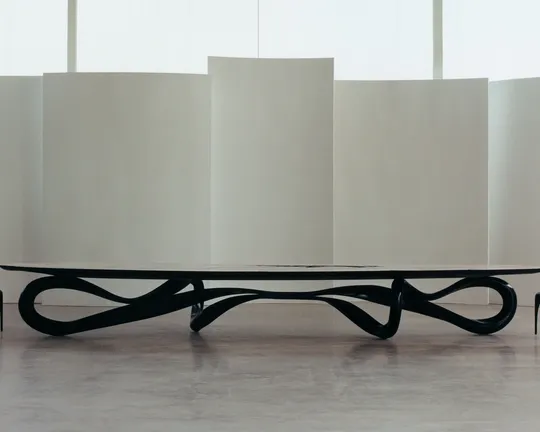 Long black sculptural table with intertwined base in a minimalist gallery, flanked by two small chairs, against curved white panels.