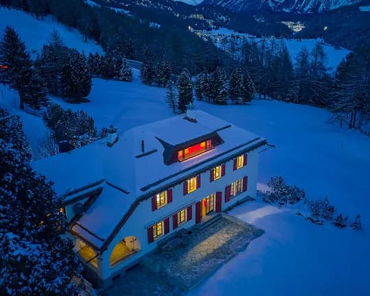 An aerial view of a snow-covered mountain hut surrounded by fir trees, a winding road and snow-covered hills beneath a bright blue sky.