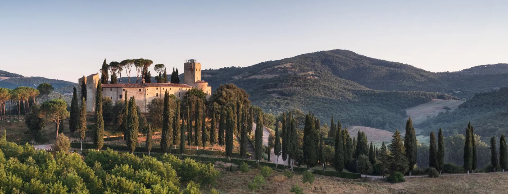 Sunlit hilltop medieval castle with a tower among tall cypress trees, overlooking rolling green hills under a clear blue sky.