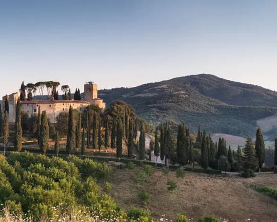 A sun-drenched medieval castle on a hill, with a tower set amongst tall cypress trees, overlooking rolling green hills beneath a brilliant blue sky.