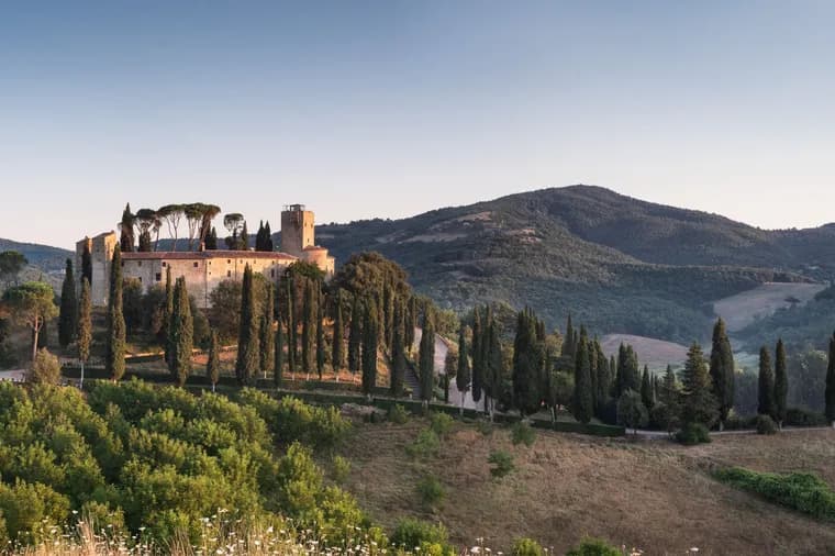 Sunlit hilltop medieval castle with a tower among tall cypress trees, overlooking rolling green hills under a clear blue sky.