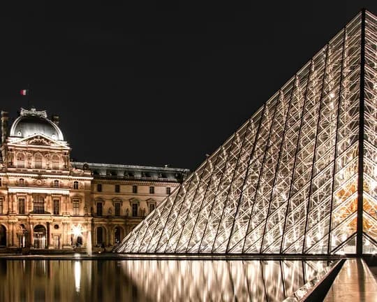 Night view of the illuminated Louvre Pyramid beside the historic Louvre palace, its glass facets reflecting in the water.