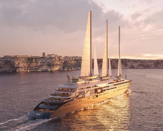 Luxurious multi-masted yacht glides along calm water beside a rocky coastline at sunset, bathed in golden light.