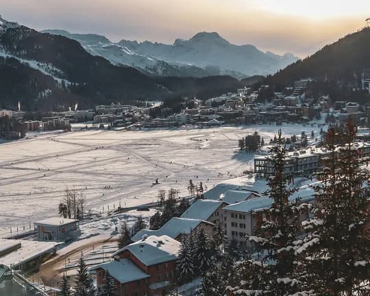 Snow-covered alpine town in a valley, ski tracks across a frozen field, with forested hills and distant snow-capped mountains at sunset.