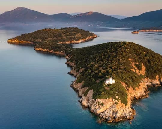 Aerial view of a forested island with rocky cliffs, a small white building at the edge, calm blue water, and distant mountains under a pastel sky.