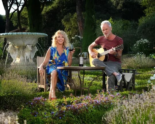 Couple in a lush garden; man plays guitar while woman sips wine at a table beside a stone fountain.