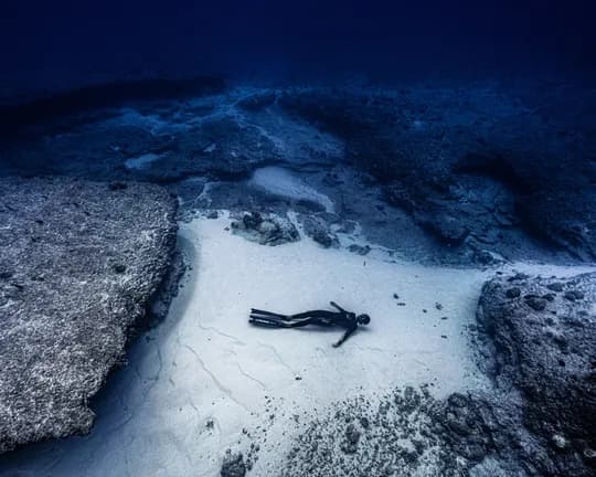 Underwater rocky seabed with a lone diver resting on pale sand between large rocks, deep blue water above.