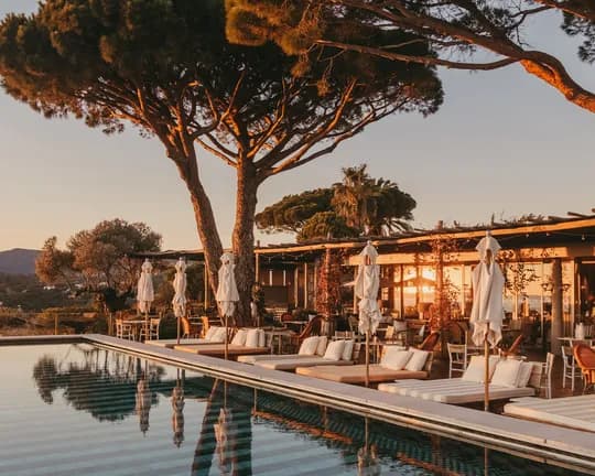 Sunset-lit resort poolside with wicker lounge chairs, white cushions, closed umbrellas, and tall trees reflecting in calm water, hills in distance.