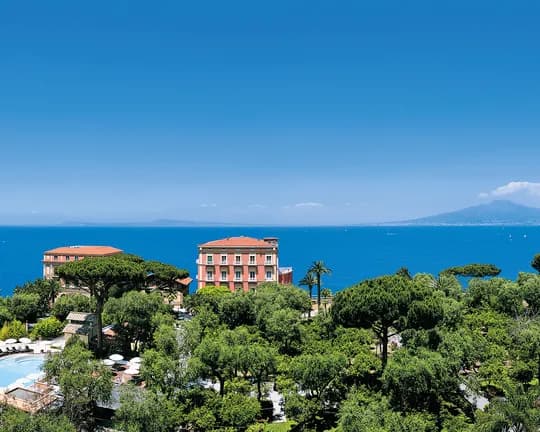 Panoramic coastal scene: pool and lounge chairs in a lush garden, red-roofed buildings, blue sea, and a distant volcano under a clear sky.