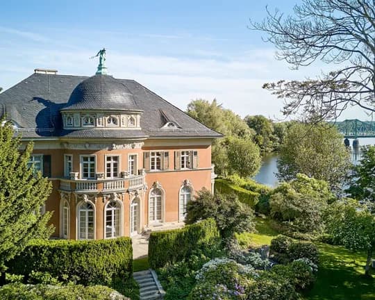 An elegant orange-coloured mansion with a domed tower, ornate windows, a statue on the roof and a lush garden on the riverbank; a bridge can be seen in the distance in the background.