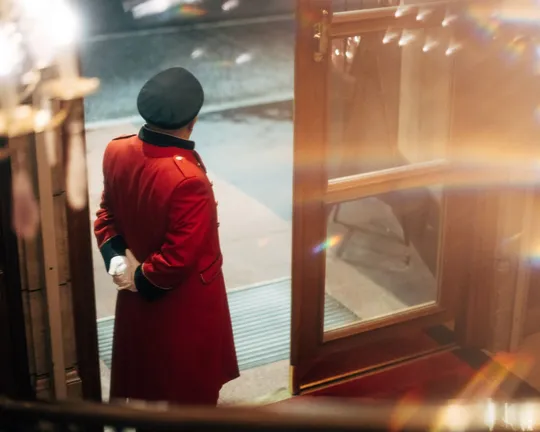 Un fattorino d'albergo con un cappotto rosso e un cappello nero è in piedi davanti a una porta a vetri aperta, con le mani incrociate dietro la schiena, e guarda fuori verso una strada delicatamente illuminata.