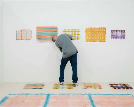 A person in a gray shirt and jeans bends over to view colorful rectangular artworks on a white gallery wall, with blue and peach floor rugs.