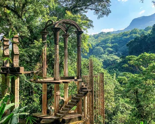 Ancient stone arches and pillars form a ruined terrace in lush jungle, overlooking verdant hills and distant mountains.