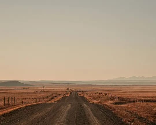 A gravel road winds through dry plains lined with fences on both sides, disappearing toward the distant hills as the sun sets.