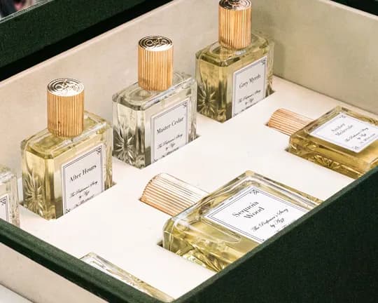 Eine Reihe von Parfümflakons aus Glas mit goldgerippten Verschlüssen in einer Vitrine; auf den Etiketten steht „Master Cedar“, „Grey Myrrh“ und „After Hours“.