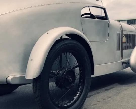 Vintage silver race car with large rounded fenders, wire-spoke wheels, and an open cockpit parked on a paved surface.