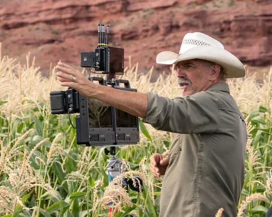 An older man wearing a beige shirt and white cowboy hat operates a large film camera rig in a cornfield, with tall stalks and a red rock cliff behind.