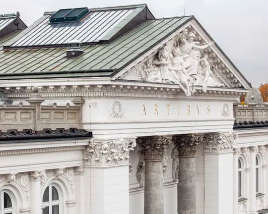 An elegant, classical-style white building with a sculptural pediment, ornate columns and golden ‘ARTIBVS’ lettering; autumnal trees in the background.