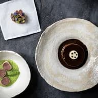 Top-down view on a dark surface: a napkin with a tiny flower-topped cake, a white bowl of green sauce with meat slices, and a beige stone plate with chocolate dessert.
