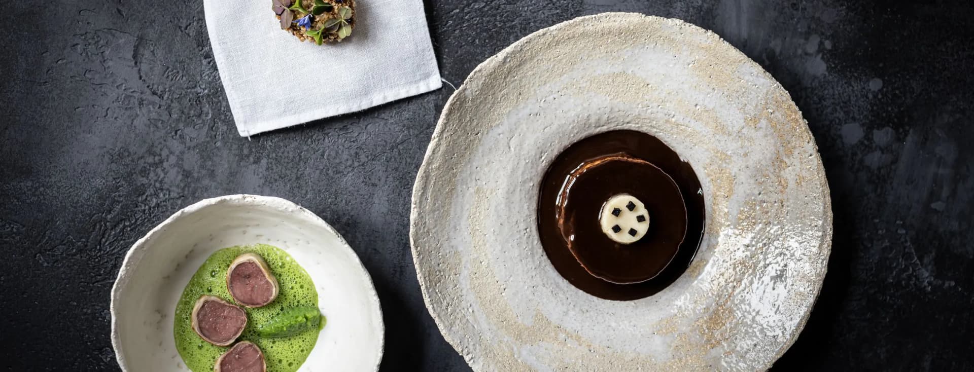 Top-down view on a dark surface: a napkin with a tiny flower-topped cake, a white bowl of green sauce with meat slices, and a beige stone plate with chocolate dessert.
