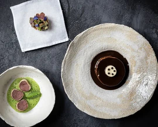 Dark surface with a pale plate holding a chocolate pool topped by a white four-hole button; a white bowl contains green sauce with pink slices; a napkin bears a floral cluster.