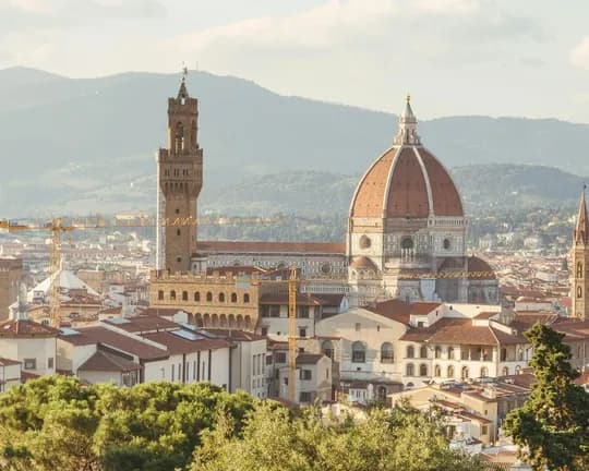 Panoramic view of Florence: the Duomo with its red dome and the Palazzo Vecchio tower amid tiled rooftops and hills in the background.