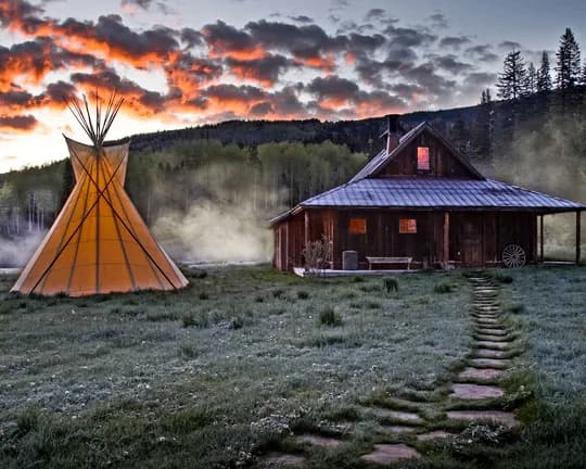 Sunset scene with a yellow tipi on the left and a wooden cabin on the right, foggy meadow, rising smoke, pine forest, and a stone path.