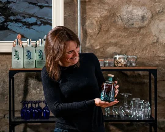 Smiling woman in a black sweater holds a bottle in a rustic room, standing before shelves of glassware and labeled bottles against a stone wall.
