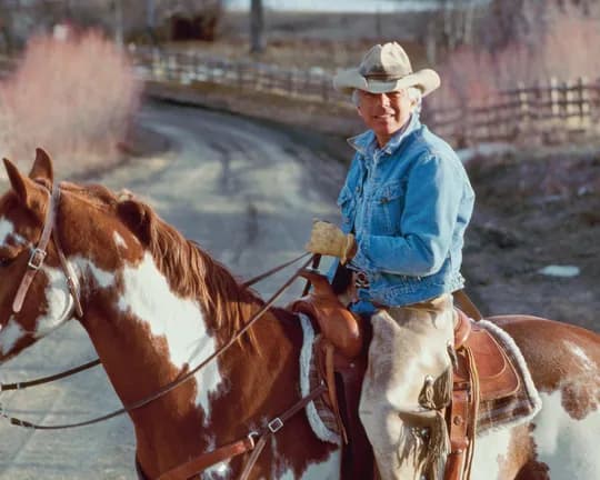 A cowboy in a light-colored hat and denim shirt rides a brown-and-white paint horse along a rural dirt road bordered by fences and bare trees.