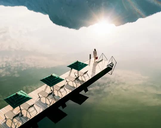 A wooden dock with green canopies and chairs extends into calm water beneath a rocky overhang; a person stands at the end as the sun shines overhead.
