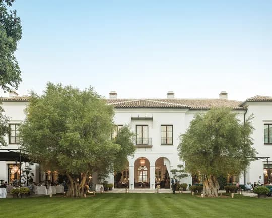 White Mediterranean-style building with tiled roof and arches, surrounded by tall trees; guests dine at tables in a shaded courtyard.