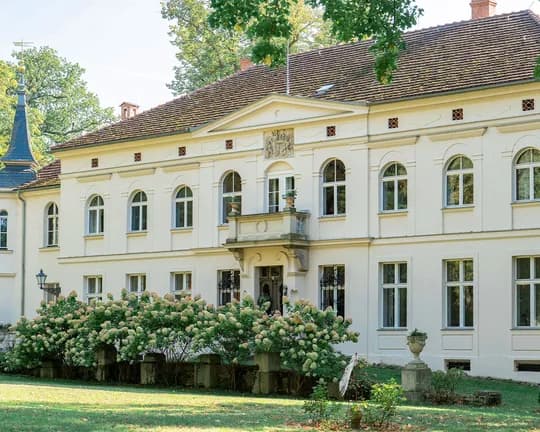 Cream neoclassical building with arched windows and a tiled roof, set in a garden with hydrangeas and trees.