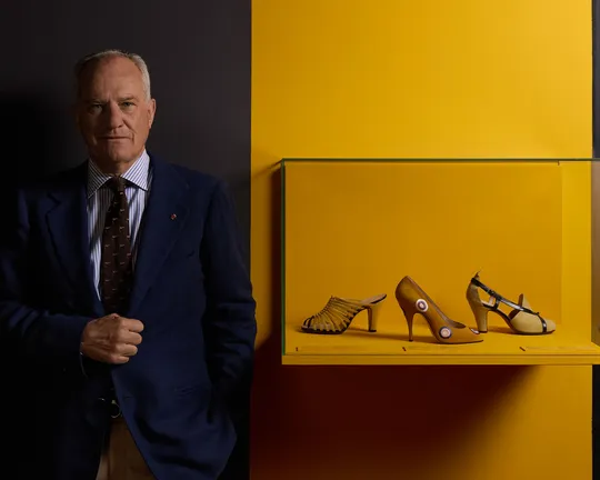 An older man in a blue suit stands beside a yellow display shelf showing three pairs of shoes against a dark backdrop.