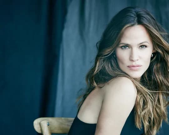 Portrait of a woman with long wavy brown hair wearing a black off-shoulder top, seated with a wooden chair behind her against a blue backdrop.