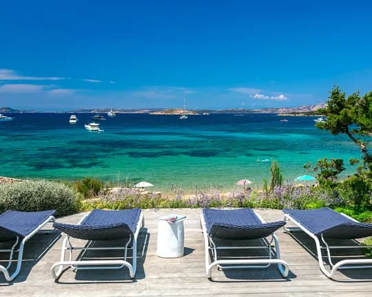 Row of blue lounge chairs on a wooden deck overlooking a turquoise sea with moored boats and distant islands under a clear blue sky.