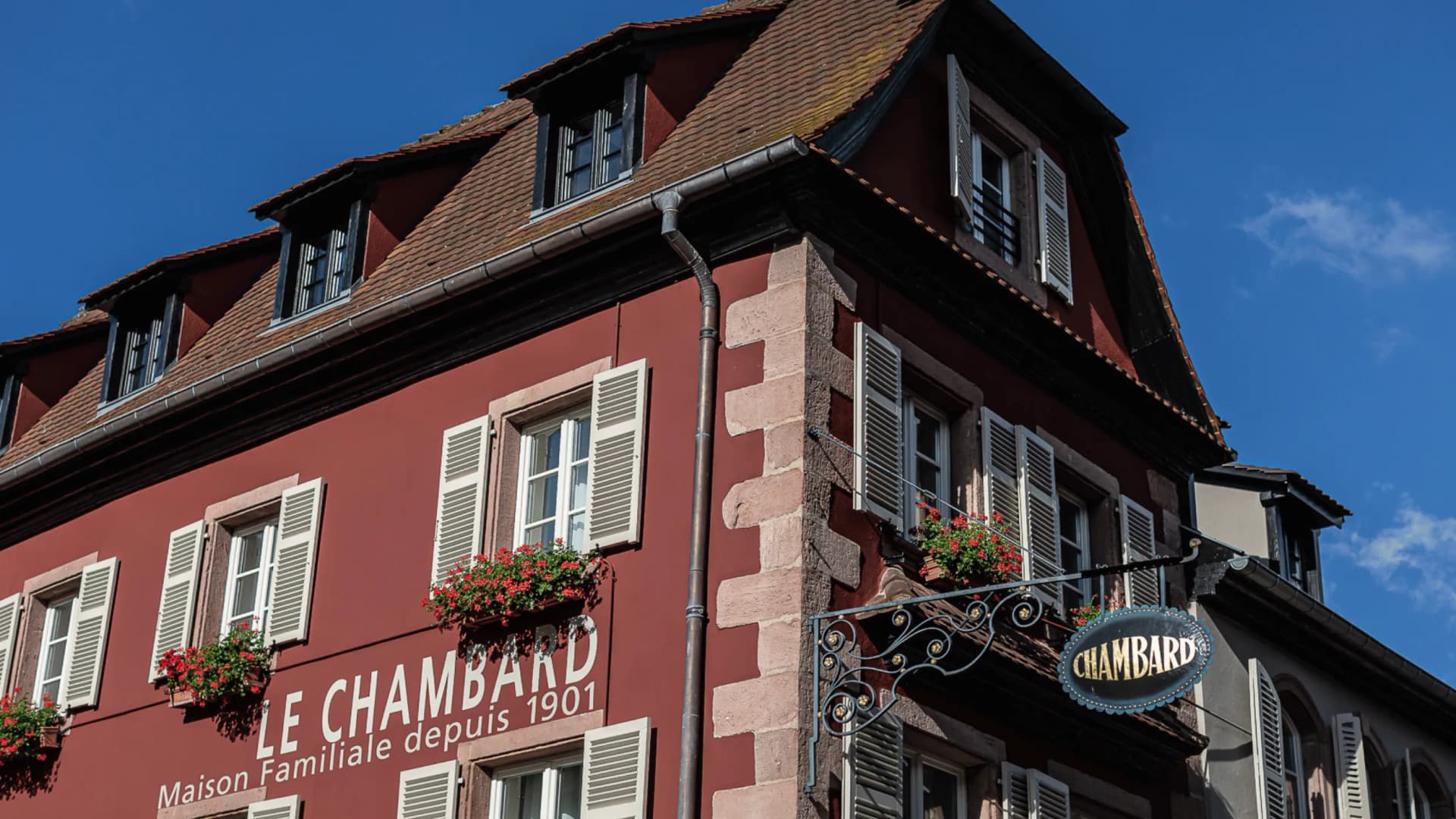 Red building with white shutters, flower boxes, and an ornate wrought-iron sign beneath a steep tile roof against a blue sky.